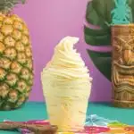 Clear cup of Dole Whip with a pineapple and a tiki glass in the background