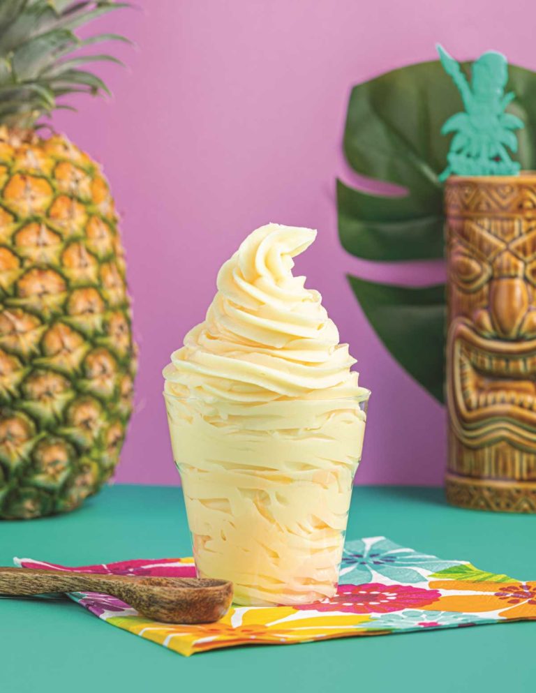 Clear cup of Dole Whip with a pineapple and a tiki glass in the background