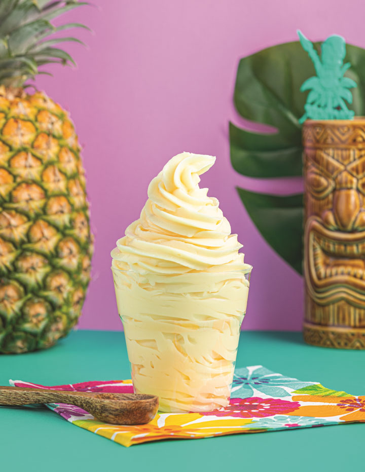 Dole whip in a cup on a colorful napkin with a whole pineapple and a tiki glass in the background